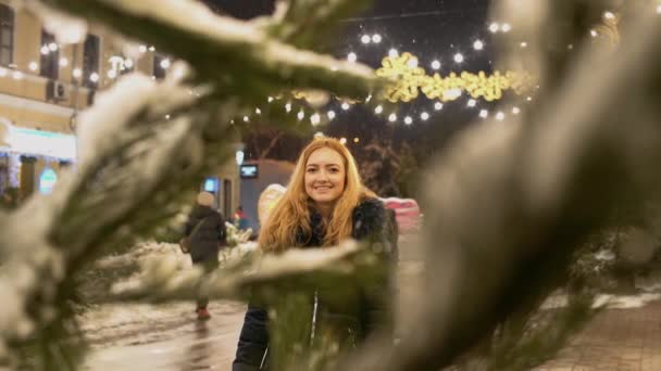 若い女性はカメラで見て近づく。フォア グラウンドで雪の中のクリスマス ツリーの枝 — ストック動画