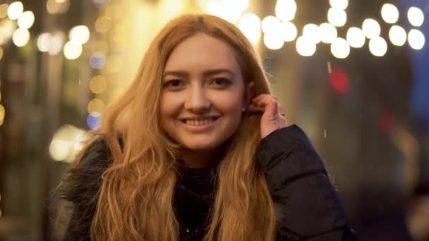 Retrato de una joven y alegre mujer sonriente mirando a la cámara al aire libre. Linda chica en el fondo de luces intermitentes de una calle festiva de invierno — Vídeo de stock