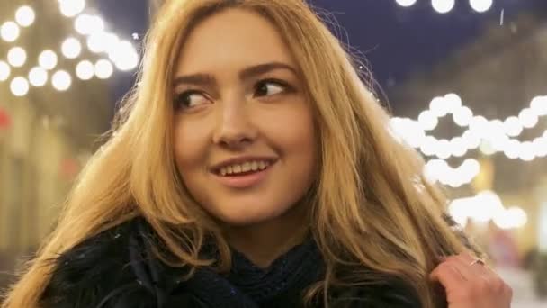 美丽的微笑的女孩在冬季夹克站在一个闪烁的花环在冬季街道上的背景下, 揉她的手掌. — 图库视频影像