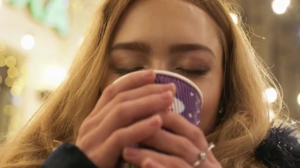 Portrait d'une jolie jeune fille avec de longs cheveux blonds debout dans une veste d'hiver dans une rue d'hiver et buvant du thé chaud dans une tasse en carton . — Video