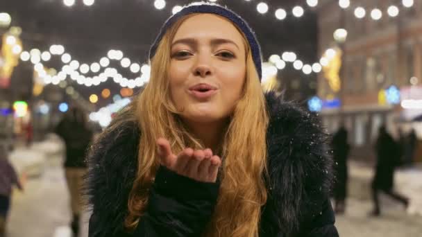 Портрет милої молодої дівчини з довгим світлим волоссям, що стоїть в зимовій куртці на святковій міській вулиці з яскравими обличчями і дме поцілунок — стокове відео