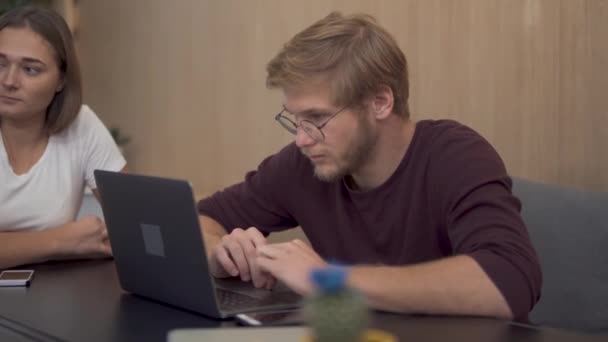 En ung skäggig kille sitter på en bärbar dator noga lyssna till den finansiella rapporten. Affärsmän möte på styrelserummet tabell. — Stockvideo