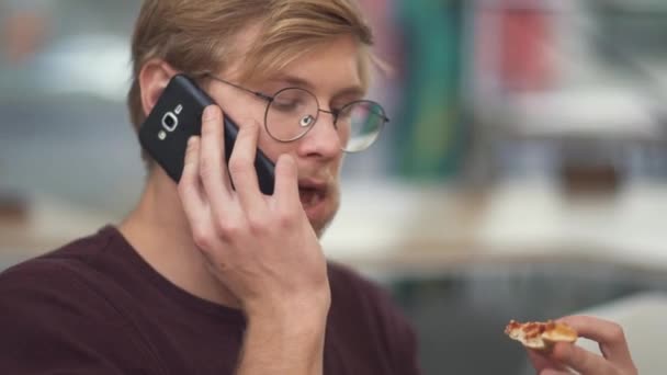 Portrét mladé vousatý chlapík s brýlemi jíst sendviče a mluví o mobilní telefon — Stock video