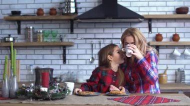 Mom and little daughter in plaid shirts have breakfast in the kitchen eating cookies and a drinking tea together