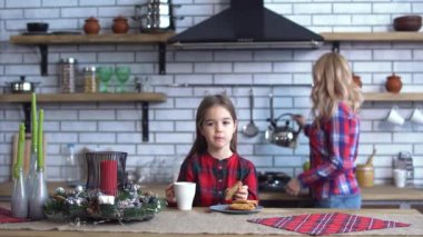 Little beautiful girl with long brown hair eating cookies with tea on the kitchen table and young mother make coffee