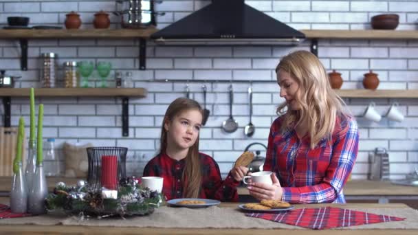 Madre con el pelo largo blong sentado en la mesa de la cocina con su hermosa hija que alimentan a la mamá con galletas — Vídeos de Stock