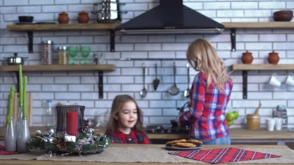 Joven madre en camisa a cuadros con pelo largo y rubio cocinando el desayuno y pequeña niña traviesa morder una galleta — Vídeos de Stock