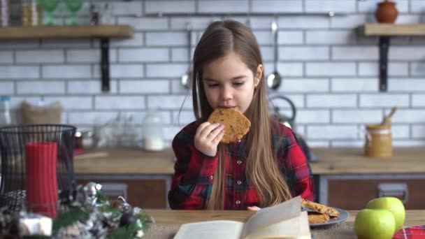 Kicsi szép lány ül a konyhaasztalon, enni a cookie-kat és leafing keresztül könyv — Stock videók