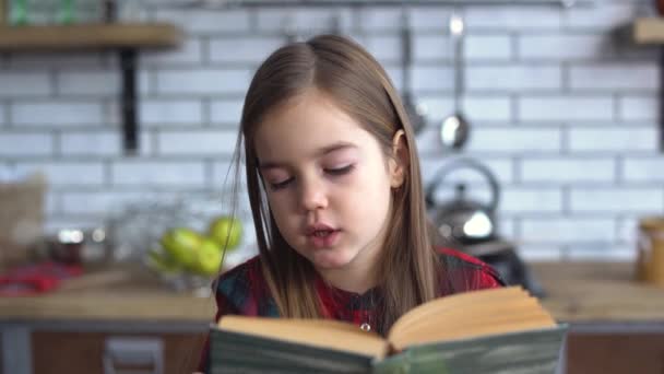 Retrato de una hermosa niña con una camisa a cuadros leyendo un libro sentado en la mesa de la cocina — Vídeo de stock