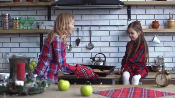 Pieni söpö tyttö istua keittiön päällä ja nuori vaalea äiti leipoa evästeet ja poista ne uunista — kuvapankkivideo