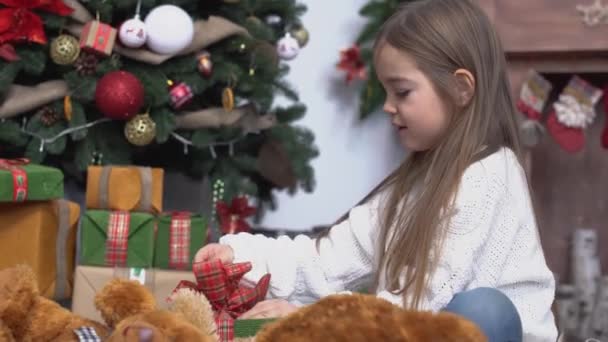 Pequena menina bonito com cabelos castanhos longos sentado perto da árvore de Natal e presentes abertos — Vídeo de Stock
