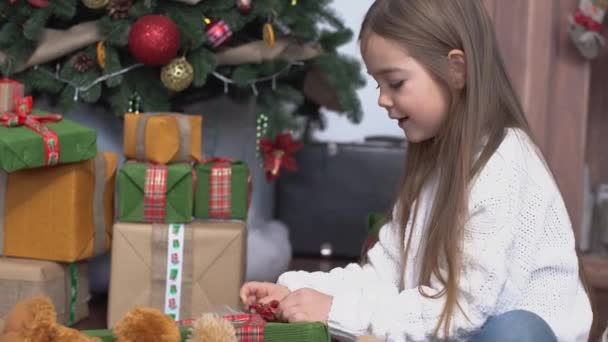 Pequena menina bonito com cabelos castanhos longos sentado perto da árvore de Natal e presentes abertos — Vídeo de Stock