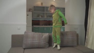 Cute jouful boy in green pajamas jumps on the sofa at home. Child grabs pillow and throws in on the floor. Happy cheerful kid. Slow motion.
