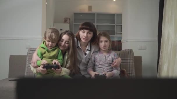 Família feliz de quatro membros jogar o jogo de vídeo. Duas crianças pequenas seguram joysticks. Férias familiares divertidas . — Vídeo de Stock