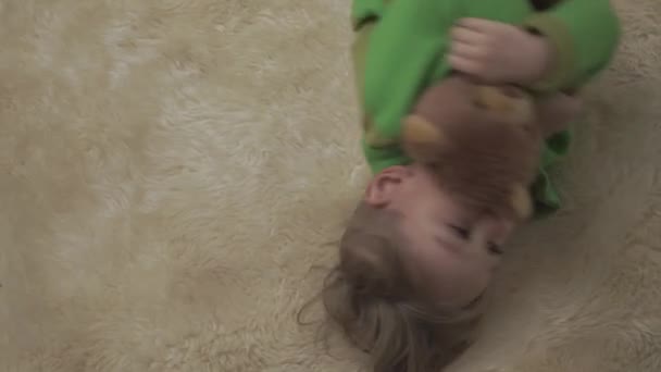 Mignon petit garçon en pyjama vert roulant sur le sol avec tapis moelleux étreignant ours en peluche. Joyeux enfant joyeux . — Video