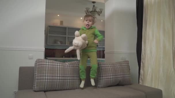 緑パジャマでかわいい小さな男の子は 自宅のソファにジャンプします 遊び心のある子は 枕を抱え 床の上でスローします 楽しいだけで自宅に喜んでいる子供 ブロンド少年のレジャー — ストック動画