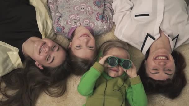 Family portrait of joyful older sisters and younger boy and girl lying on the carpet in the room. Funny little boy in pajamas looks through binoculars causing laughter and joy to his relatives. Fun — Stockvideo