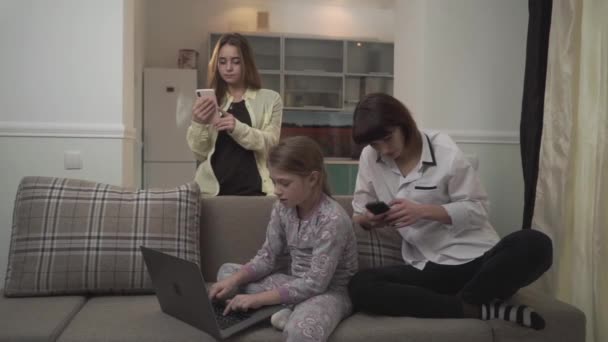 Two older sisters texting messages on cell phones younger girl typing on laptop sitting on the couch in the room. Family spends time together but not pay attention to each other. — 비디오