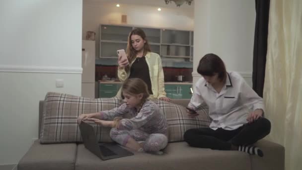 Dwie starsze wiadomości SMS-y siostry na telefony komórkowe młodsza dziewczyna wpisując na laptopie siedzi na kanapie w pokoju. Mała dziewczynka blisko laptopa i przytula jej siostra. Dziecko potrzebuje więcej miłości z jej rodziny — Wideo stockowe