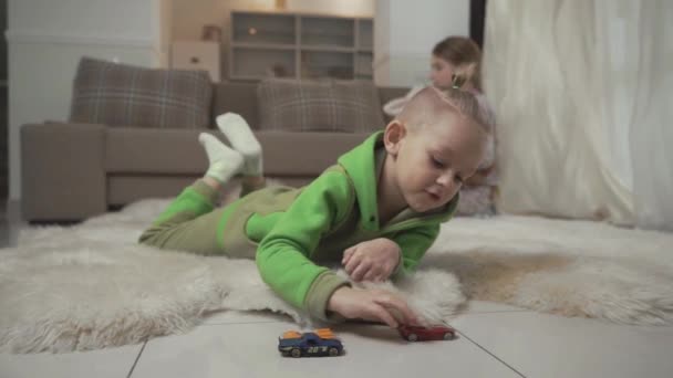 Little boy with stylish haircut plays with his toycars lying on the floor on fluffy carpet. Sister playing with a teddy bear on the background. — Stock video