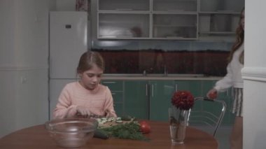 Younger sister helps the older sister coocking vegetable salad for breakfast sitting at a small table in the cozy kitchen. Sisters relationship.