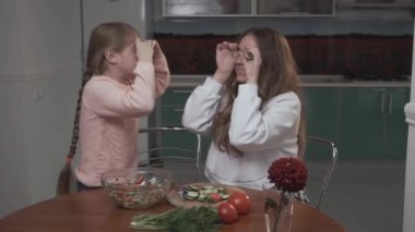 Joyful sisters fooling around coocking a vegetable salad applying cucumber slices to their eyes sitting in the kitchen. Sisters relationship.