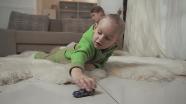 Lille dreng med stilfuld frisure leger med sine legetøjsbiler liggende på gulvet på fluffy tæppe. Søster leger med en bamse på baggrunden . – Stock-video