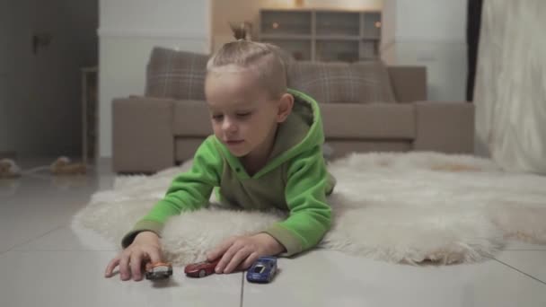 Porträtt av pojke med snygg frisyr leker med toycars liggande på golvet på fluffig matta. — Stockvideo