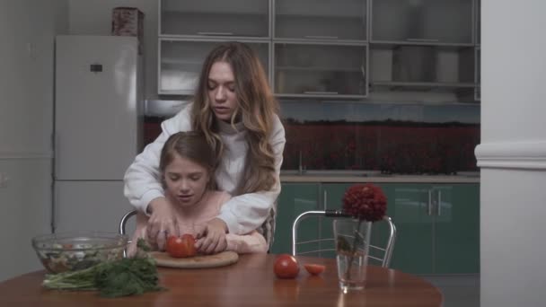 Older sister teaches younger sister to cut tomatoes for salad. Two sisters cocking vegetable salad while sitting at the table in the kitchen. Sisters relationship. — Stockvideo