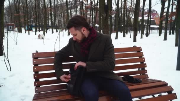 Ein bärtiger Mann sitzt auf dem Bankett im Winterpark. Geschäftsmann öffnet seinen Koffer und bekommt Buch — Stockvideo