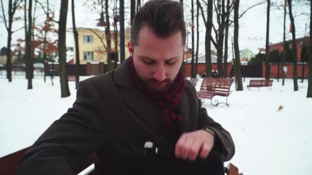 Elegante uomo d'affari seduto su una panchina in un parco invernale disegnando una tavoletta con una valigetta. Bello giovane uomo che lavora su tablet computer all'aperto — Video Stock