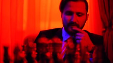 Bulanık bir arka plan üzerinde oturan iş elbiseli düşünceli işadamı satranç oynar. Parlak kırmızı ışıkta çekim.