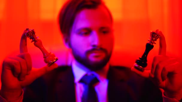 Retrato de um empresário de sucesso em um terno de negócios segurando uma figura de xadrez branco e preto em seus braços sentados na poltrona em luz vermelha brilhante. Oportunidade de escolher em suas mãos — Vídeo de Stock