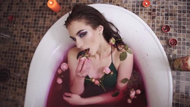 Härlig kvinna tar ett bad med rosor i spa. Vacker dam sätter långsamt ros i munnen letar upp i kameran. Söt flicka avkopplande i badrummet med brinnande ljus. — Stockvideo