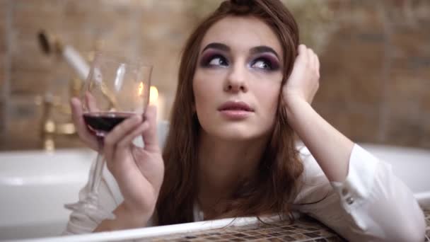 Мрійлива молода жінка в білій сорочці п'є червоне вино з високого келиху, сидячи в розкішній ванні і посміхаючись крупним планом . — стокове відео