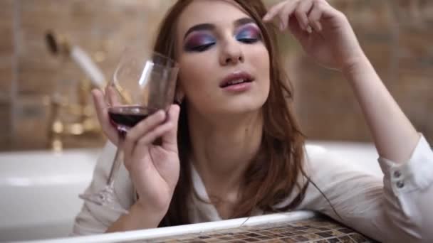 Retrato de una chica pensativa tomando un baño con maquillaje brillante en una camisa blanca con copa de vino — Vídeos de Stock