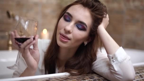 Porträt eines verspielten Mädchens, das mit hellem Make-up in einem weißen Hemd mit Weinglas baden geht. sinnliche Mädchen genießen in der Badewanne — Stockvideo