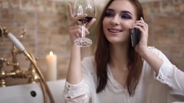 Portrait femme parler par téléphone portable faire des gestes avec verre de vin dans la salle de bain gros plan — Video