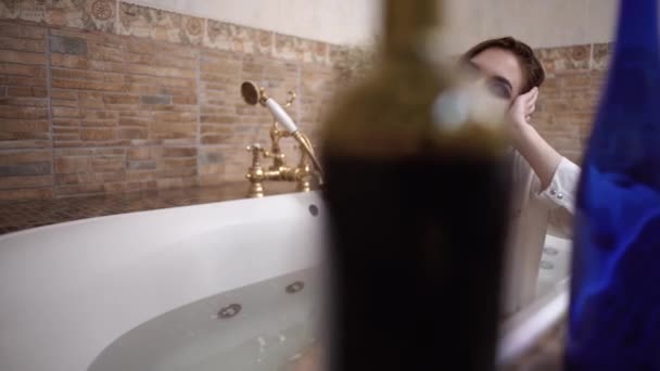 Ritratto di giovane donna sconvolta che fa un bagno con trucco luminoso in una camicia bianca con bicchiere di vino. Due bottiglie in primo piano sul bordo del bagno . — Video Stock