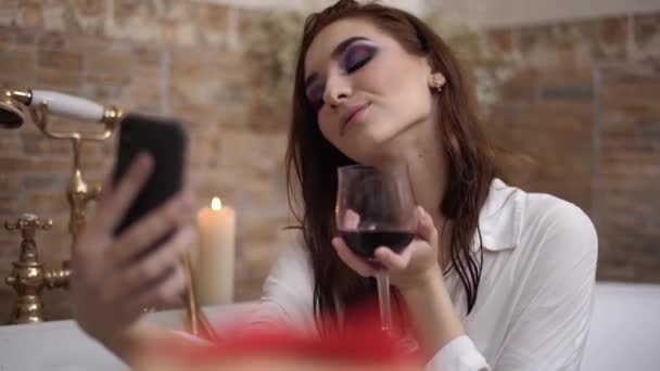 Jovem com maquiagem brilhante fazendo selfie sentado no banho com copo de vinho tinto. Menina bonito relaxante no banheiro — Vídeo de Stock