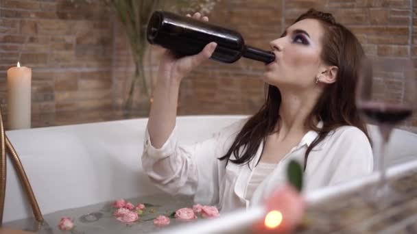 Giovane donna triste con trucco luminoso beve vino dalla bottiglia sdraiata in bagno con fiori . — Video Stock