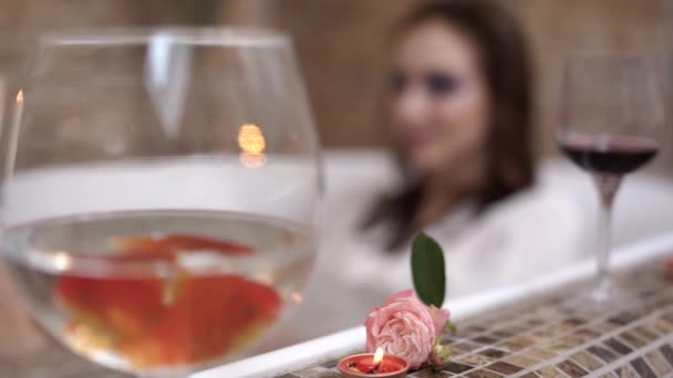 Glass with red fish are in the bath edge close up. Woman lying in bath holding small rose — Stock Video