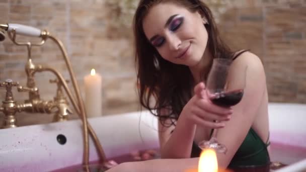 Портрет досить чуттєвої дівчини з яскравим макіяжем, що приймає ванну з трояндами і п'є вино . — стокове відео