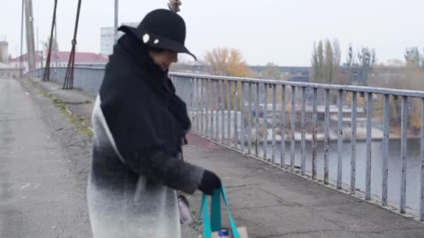 Ενήλικη γυναίκα σε ένα πλατύγυρο καπέλο βόλτες σε όλη την γέφυρα στο γκρι φυσάει κρύο και αποφάσισε να θέσει την τσάντα, το μπουκάλι και το χαρτί στην άσφαλτο — Αρχείο Βίντεο