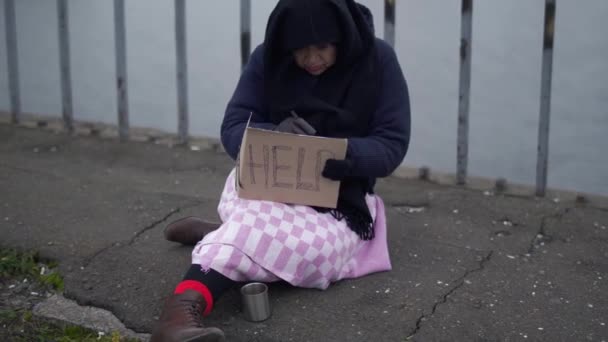 Взрослая бездомная женщина сидит на мосту в холодную ветреную серую погоду и просит милостыню, и помогает, и пишет что-то на коробке — стоковое видео