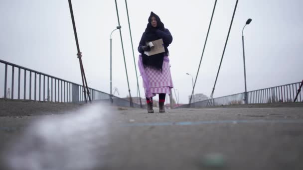 Vuxen hemlös kvinna vistelse på bron nära flodhamnen i kallt blåsigt grått väder ber om hjälp och tigger pengar — Stockvideo