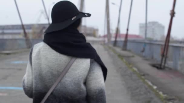Volwassen vrouw in een hoed met brede rand loopt over de brug in koude winderige grijs en zich omdraait — Stockvideo