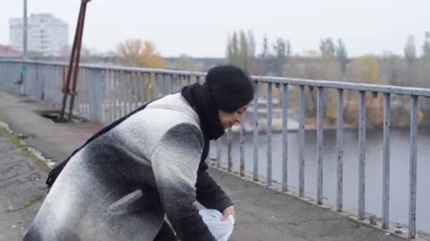 Mulher adulta com cabelo preto em pé em uma ponte em um casaco. Ela está removendo um lenço preto . — Vídeo de Stock