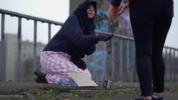 Eine ältere Obdachlose sitzt mit ausgestreckter Hand auf der Brücke bei kaltem, windigem grauen Wetter und bittet um Almosen und Hilfe, und eine Frau hält an, um Münzen zu legen..