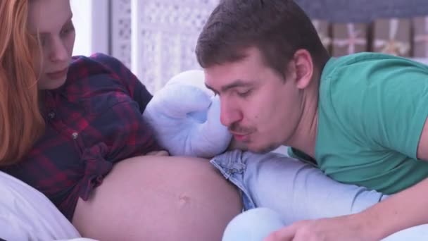 漂亮的孕妇和她的丈夫躺在床上。爸爸亲吻妻子玩软玩具的肚子。产妇概念。妊娠。幸福的家庭. — 图库视频影像